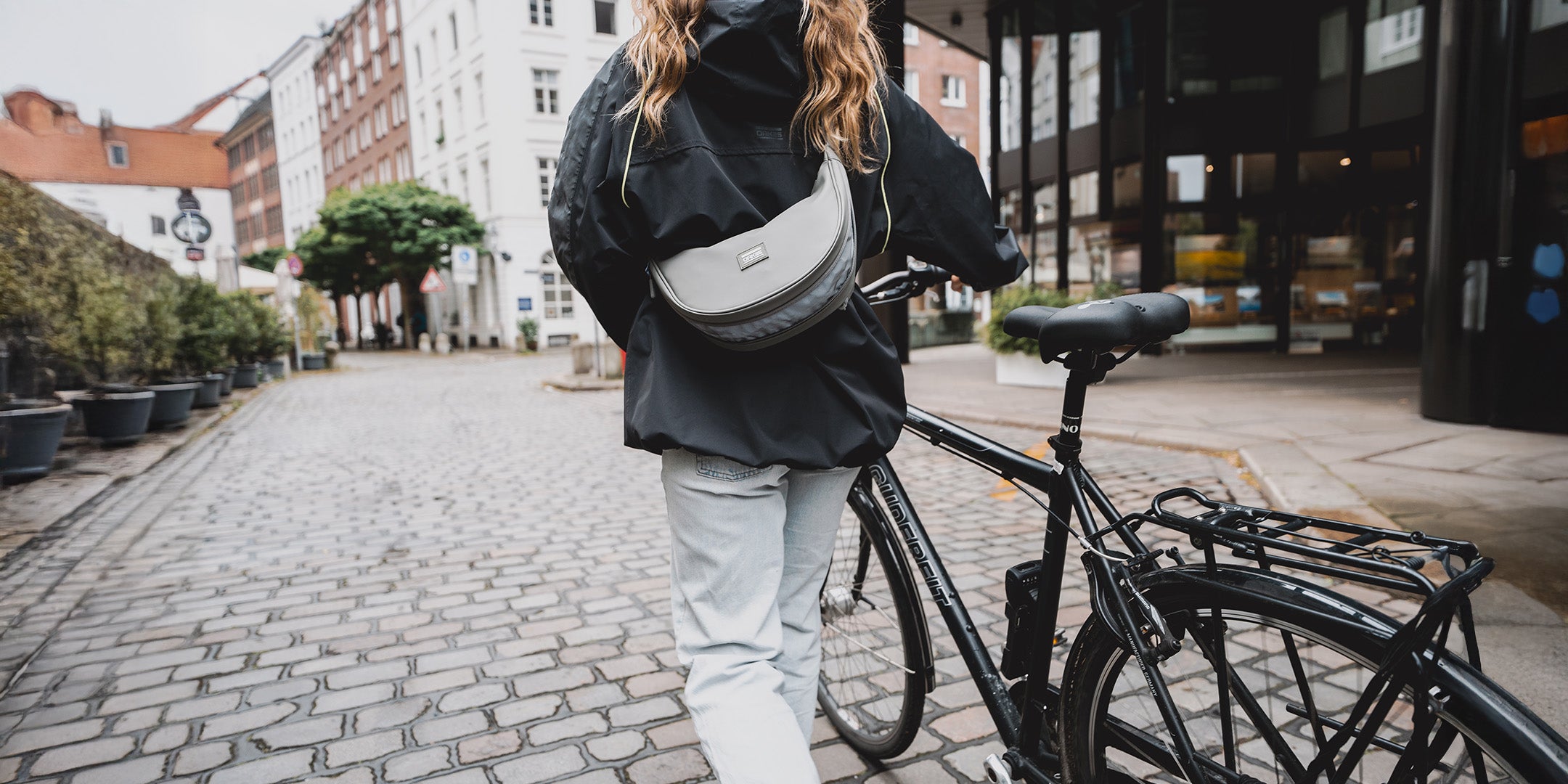 Frau in schwarzer Jacke mit grauer Halbmond-Tasche auf gepflasterter Straße neben schwarzem Fahrrad