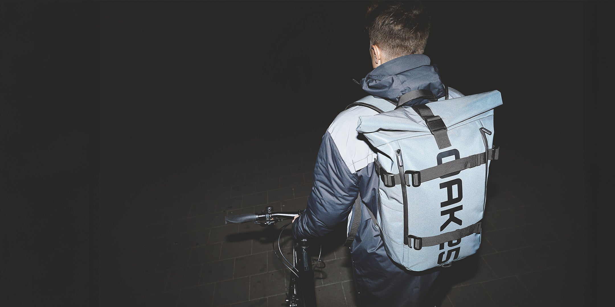 Person mit blauem Rucksack mit schwarzem Schriftzug 'OAK25' auf dem Rücken hält Fahrradlenker in dunkler Umgebung