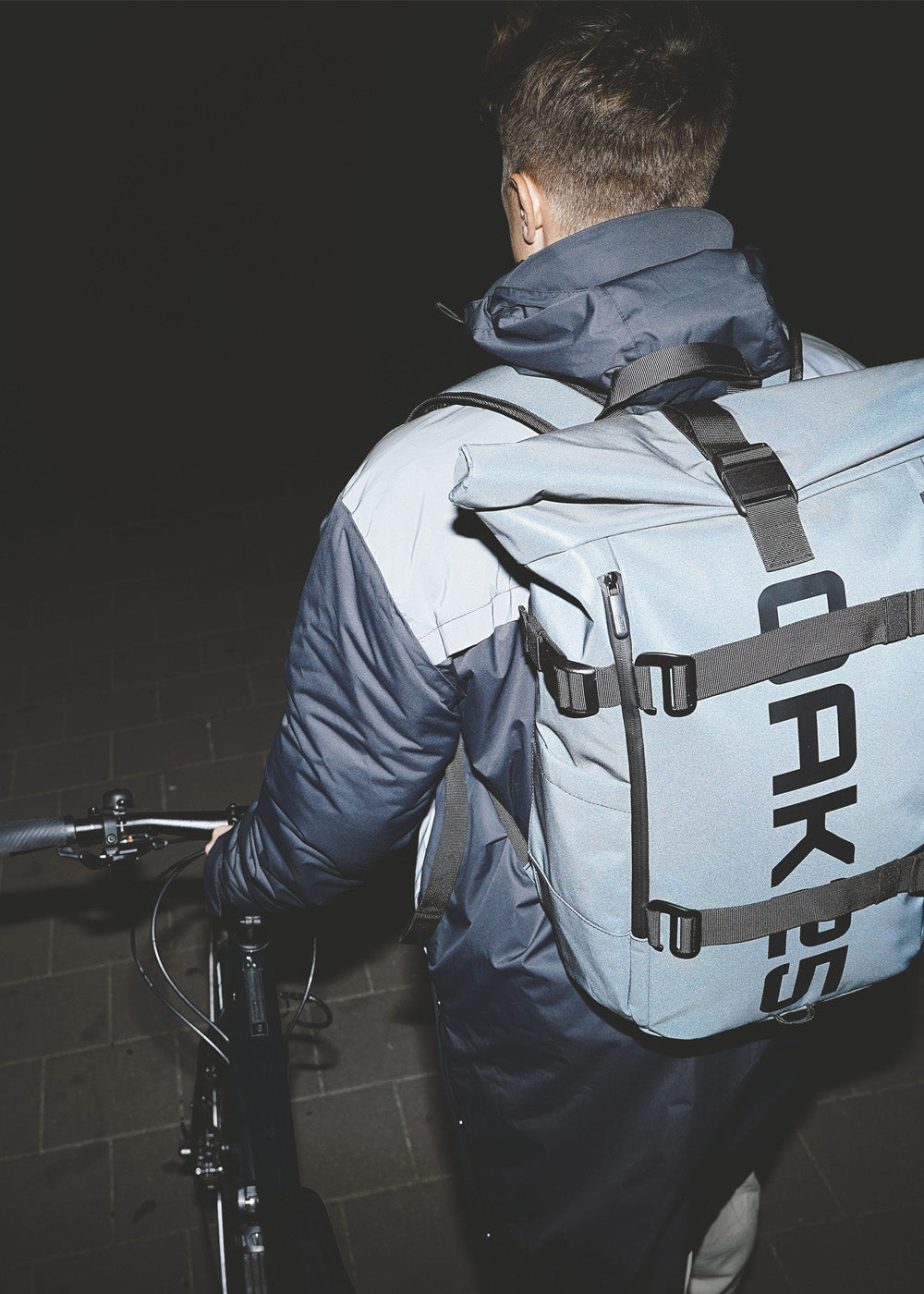 Person mit kurzem braunem Haar von hinten, trägt dunkelblauen Mantel und hellblauen Rucksack mit schwarzem Schriftzug 'OAK25 ', hält ein Fahrrad