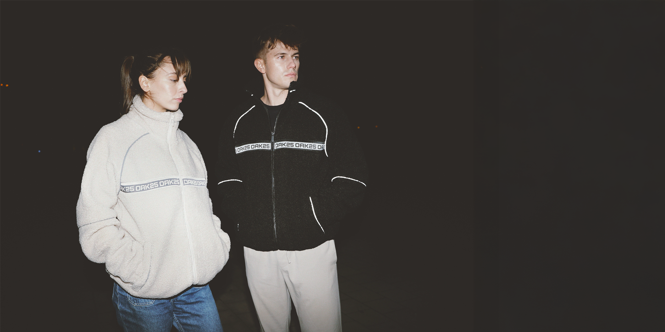 Frau und Mann tragen OAK25-Jacken, Frau weiße Fleecejacke, Mann schwarze Jacke mit Reflektoren bei Nacht